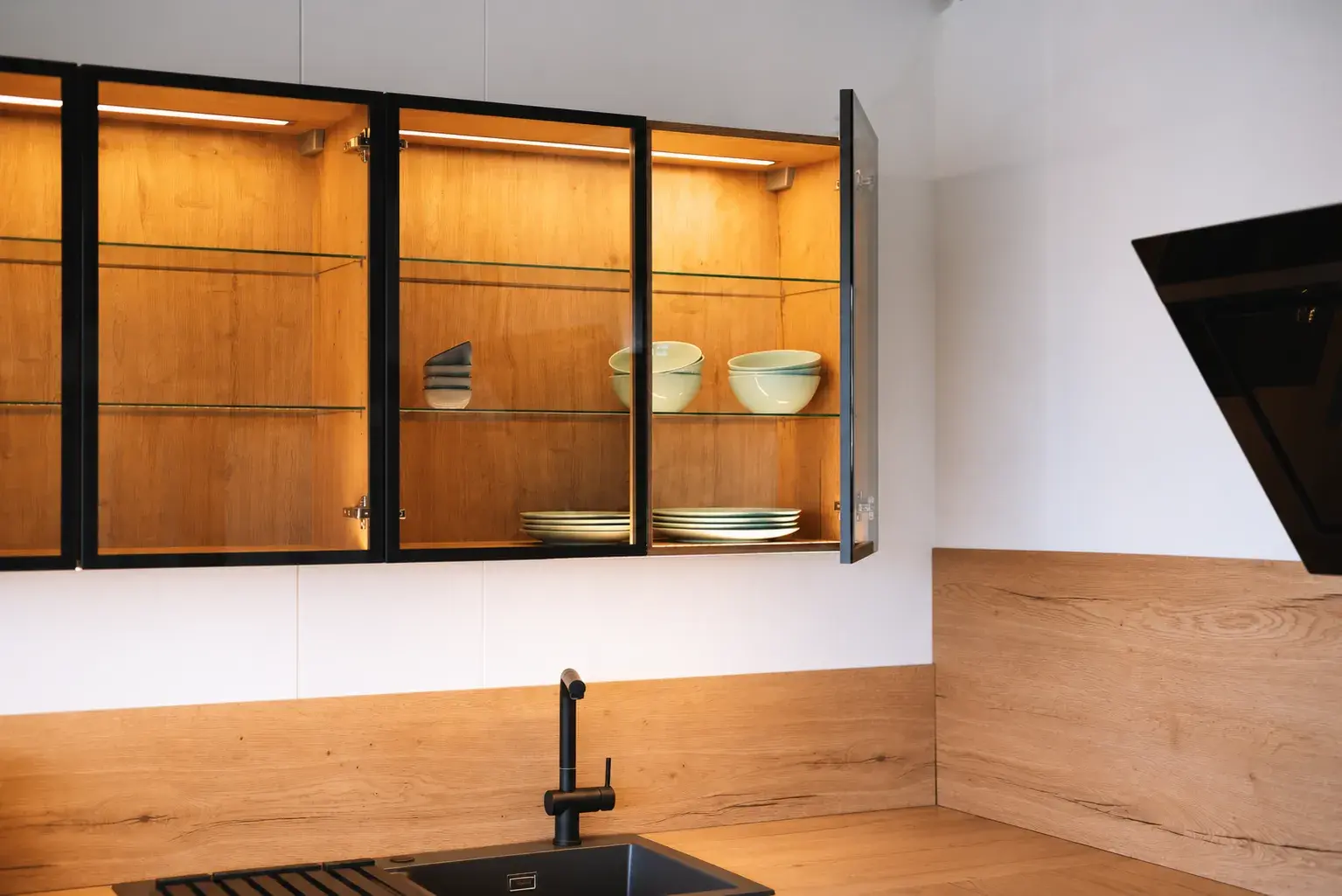 Beleuchtete Glasoberschränke mit schwarzen Rahmen, Glasböden und Holzoptik in moderner Küche mit schwarzer Spüle