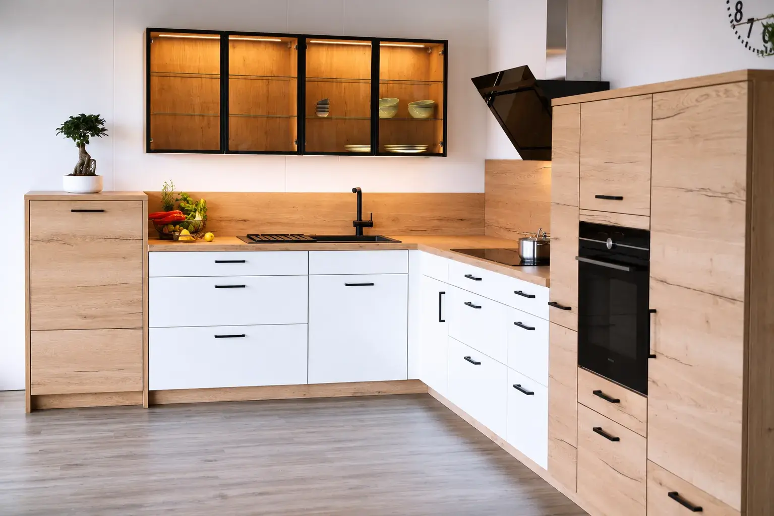 Moderne L-Küche in Weiß und Eichenholzoptik mit schwarzen Griffen, beleuchteten Glasvitrinen, Kochfeld und integriertem Backofen