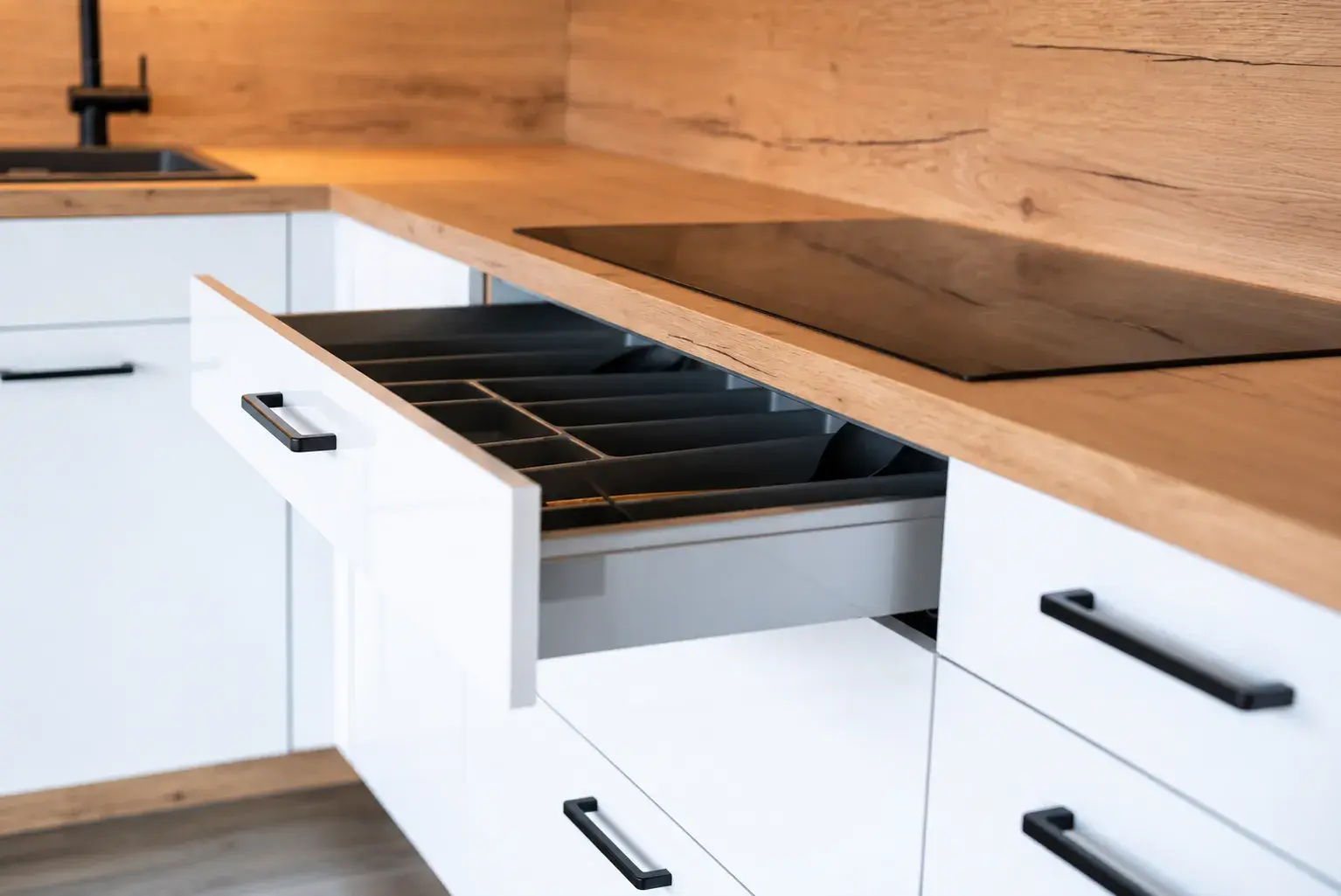 Moderne weiße Küche mit geöffneter Schublade, praktischem Besteckeinsatz, Arbeitsplatte in Holzoptik und schwarzem Kochfeld